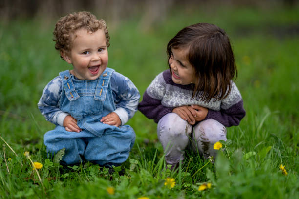 Two sweet little siblings sit in the grass together on a cool spring morning.  They are each dressed casually and are smiling at each other as they pose for the camera.
