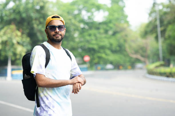 A young man wearing a yellow cap and tie-dye shirt checks his smartwatch while walking outdoors on a tree-lined path. He carries a backpack and appears focused on the device