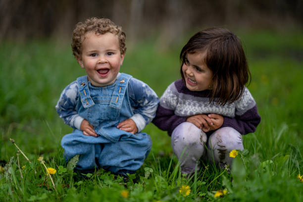 Two sweet little siblings sit in the grass together on a cool spring morning.  They are each dressed casually and are smiling at each other as they pose for the camera.
