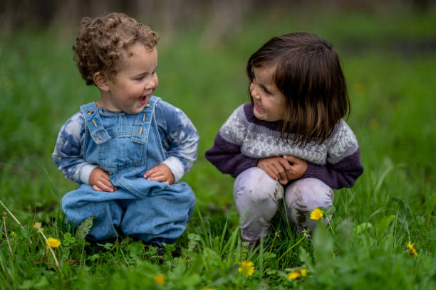 Two sweet little siblings sit in the grass together on a cool spring morning.  They are each dressed casually and are smiling at each other as they pose for the camera.