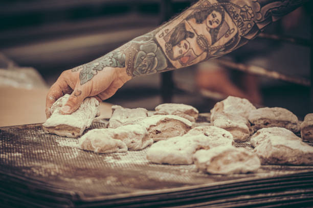 A baker with tattoos puts raw dough buns on the baking tray in the bakery