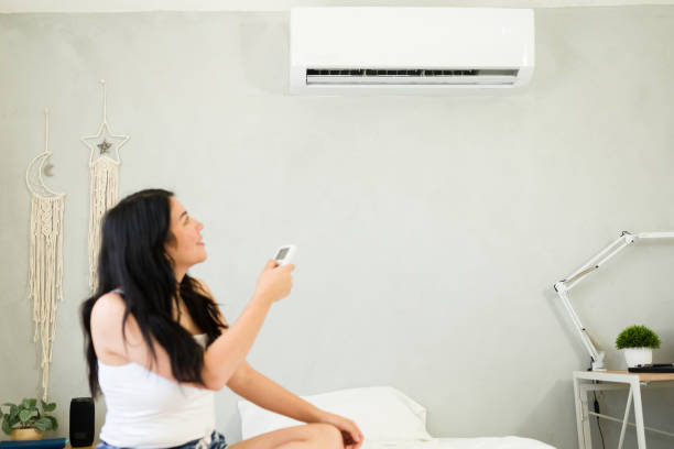 istock Young woman sitting comfortably on bed, adjusting a mini split air conditioning unit with remote control during a hot summer day. Focus on ac unit 2153468381