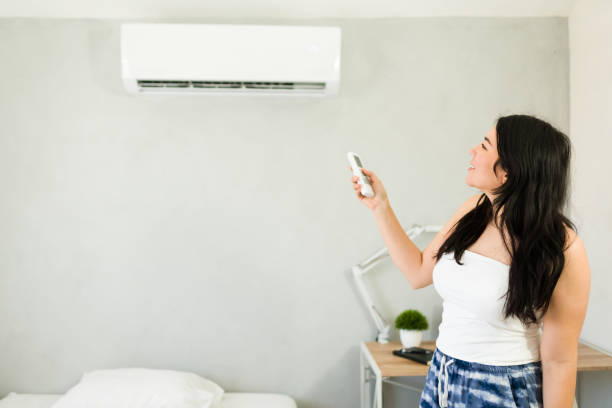 istock Happy woman adjusting mini split air conditioning unit with remote control on a hot summer day next to bed 2153468395