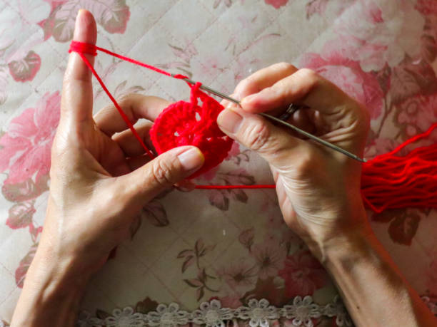 This close-up shot captures a woman's hands skillfully crocheting a piece of green yarn under the warm sunlight. The image highlights the intricate details of the crochet work, emphasizing the texture of the yarn and the precision of the craft. It evokes a feeling of relaxation and the joy of creating something handmade.