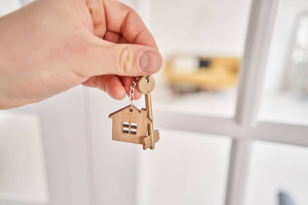 Men hand holding key with house shaped keychain. Modern light lobby interior. Mortgage concept. Real estate, moving home or renting property.