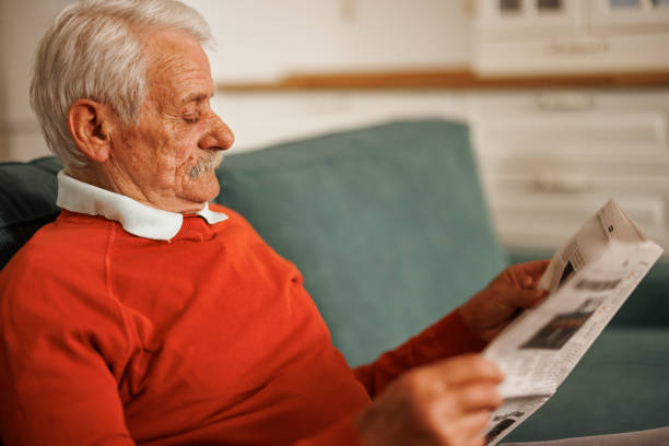 Close-up portrait of gray haired senior man sitting on sofa and reading newspaper in living room at home