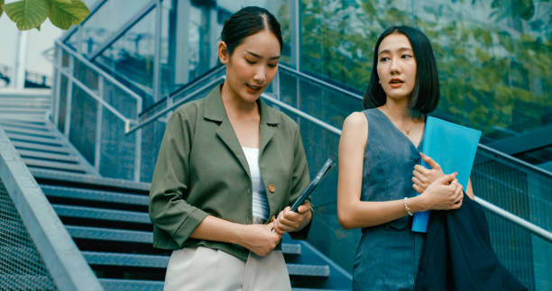 istock Two young Asian businesswomen discussing work while holds digital tablet and carries documents, walking down stairs outside office building. Sustainable business outdoor. 2153498653