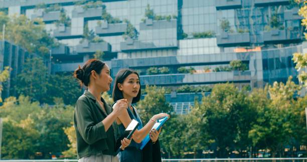 Two young Asian businesswomen discussing work while holds digital tablet and carries documents while walking outside office building. Sustainable business outdoor.