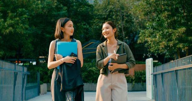 istock Two young Asian businesswomen discussing work while holds digital tablet and carries documents while walking outside office building. Sustainable business outdoor. 2153498676