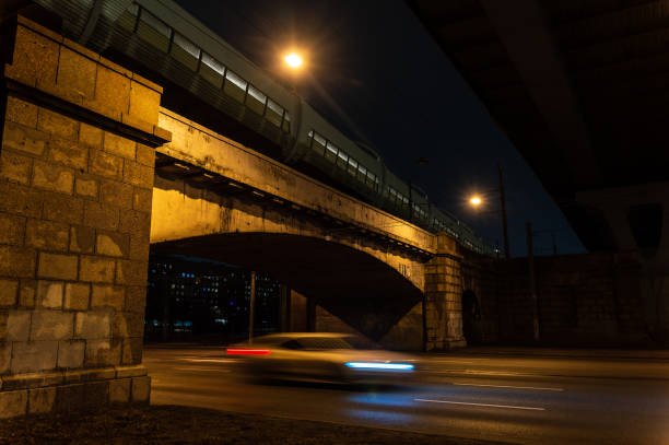 A car is driving under a bridge at night. The bridge is lit up, and the car is moving quickly. The scene is dark and moody, with the car's headlights illuminating the road ahead