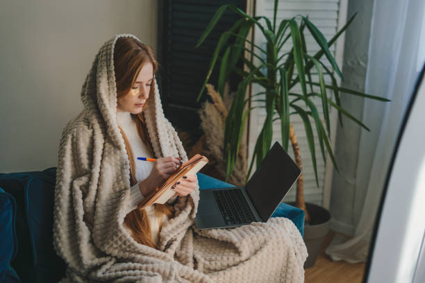 istock Image of a young woman wrapped in cozy blanket taking notes on her notebook and using her laptop computer to manage her tasks at home. 2153502889