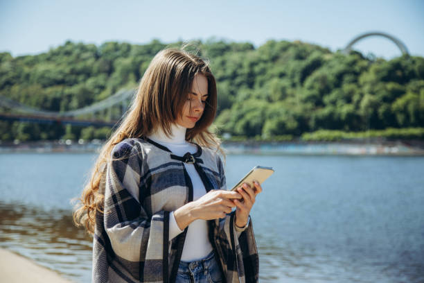 A young woman in a poncho on the bank of the river is typing a message on the phone. A young woman in a poncho on the bank of the river is typing a message on the phone. Work online anywhere. walking beach women sadness stock pictures, royalty-free photos & images