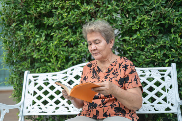 Elderly woman sits on a bench reading a book, enjoying a peaceful moment surrounded by greenery.