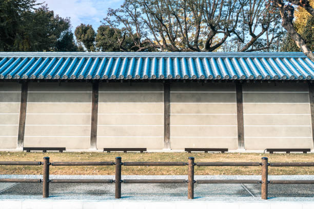 Captured in serene daylight, this image features a traditional Japanese wall with a striking blue tiled roof. The structure's simplicity and elegance are complemented by the lush greenery and trees in the background. The clean lines and muted tones of the wall provide a perfect backdrop for visual projects, allowing for ample space for overlays or text in the foreground. The image exudes a calm and peaceful ambiance, reflecting the essence of traditional Japanese architecture.