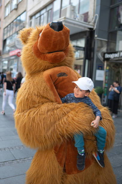A 3 year old multiracial boy is joyfully hugging a person dressed in a bear costume on a busy street, embodying warmth and happiness