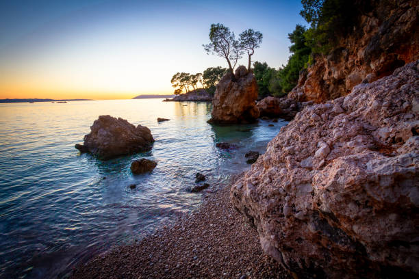 A captivating photograph of a tranquil coastal sunset over a rocky shoreline. The image captures the serene beauty of the calm ocean waters reflecting the warm hues of the setting sun. Rugged rocks and pebble-strewn shores add texture and depth to the scene, with a distant island covered in pine trees completing the picturesque view. Ideal for use in travel, nature, and landscape-themed projects, this photo evokes a sense of peace, natural beauty, and the enchanting allure of coastal sunsets.