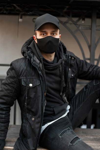 istock Handsome young man model with black cap and protective medical mask in black jacket and hoodie sits on the street 2153631628