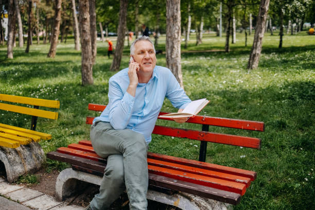 istock Man reading a book while sitting on park bench and talking on mobile phone 2153642981