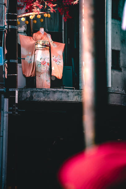 A mannequin dressed in a beautiful, traditional Japanese kimono is showcased on a balcony. The kimono features intricate floral patterns and vibrant colors. The setting appears to be an urban area with a mix of modern and traditional elements, including hanging lanterns and an aged concrete building.