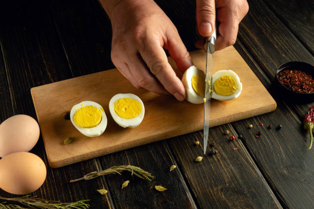 istock Slicing boiled eggs with a knife in the hands of a cook for preparing dinner on the kitchen table. Space for advertising on a dark background 2153674808