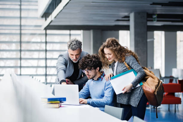 istock Handsome university professor helping students with final project, showing them research paper, book for thesis in library. 2153693097