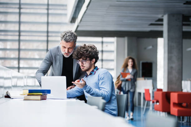 istock Handsome university professor helping student with final project, showing him research paper, book for thesis in library. 2153693204