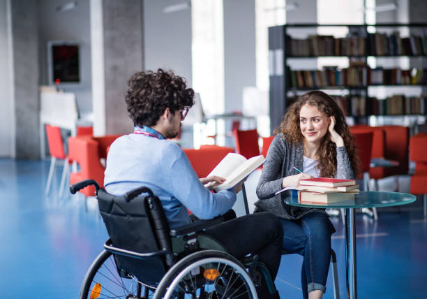 istock Handsome university student in wheelchair studying in library with classmate, preparing for final exam, presentation. Study group. Teamwork skills. 2153693413