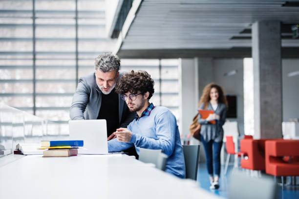 istock Handsome university professor helping student with final project, showing him research paper, book for thesis in library. 2153693597