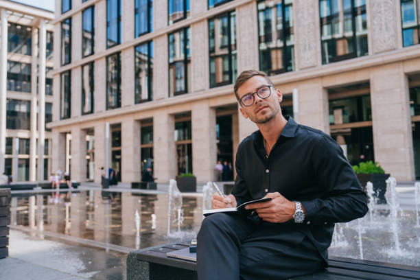 istock man in a black shirt and glasses takes notes on a notepad while sitting by a fountain in a modern urban area 2153699284