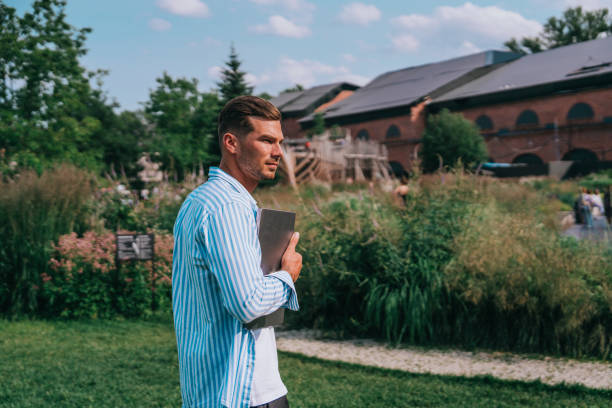 man in a striped shirt holds a laptop while standing in a park, with greenery and a red brick building in the background