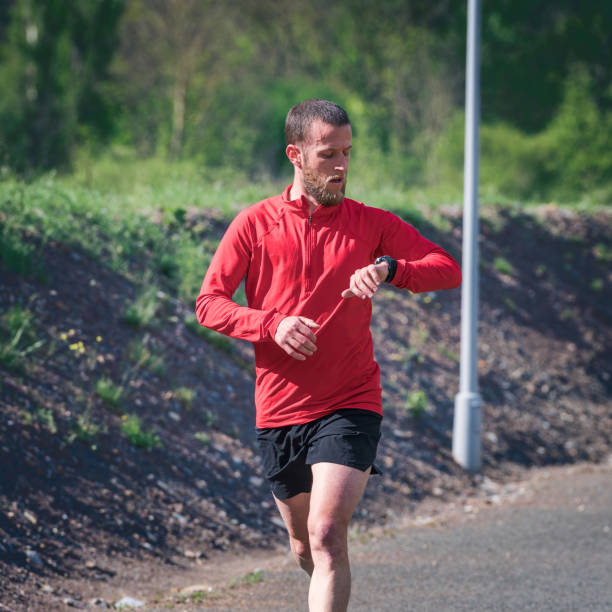 A man in a red T-shirt and black shorts runs along a path in a park. He is wearing a sports watch or wrist gps, is looking at it and is concentrating on his pace. Concept of using technology in sport.