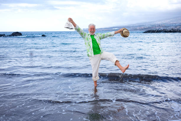 Happy cheerful elderly man barefoot on the water's edge still fully dressed while jokingly lifting one leg with open arms holding shoes and hat in hand. Peaceful retired life