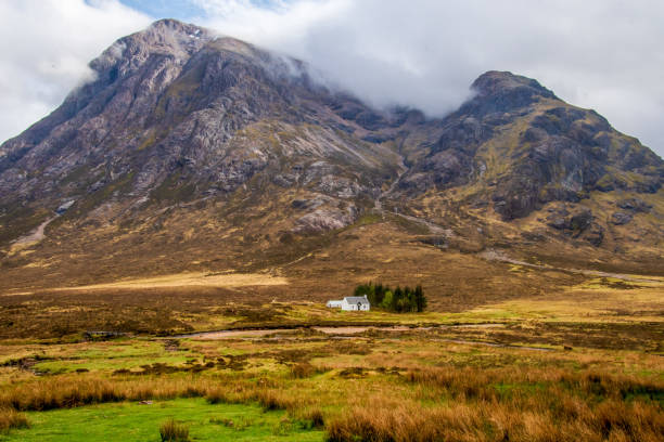 A small cottage house at the bottom of hills in  Scottish Highlands with some trees grow nearby, hills hidden in clouds, dry brown grass around, place 5 minutes from Three Sisters by car