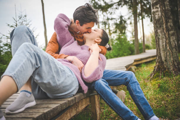 A young man and woman embrace lovingly while sitting on a park bench, with the natural scenery of fields and trees creating a serene and picturesque outdoor environment.