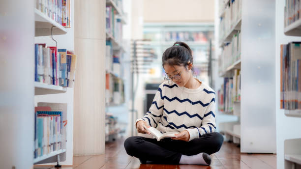 istock A girl is sitting on the floor in a library reading a book 2153750419