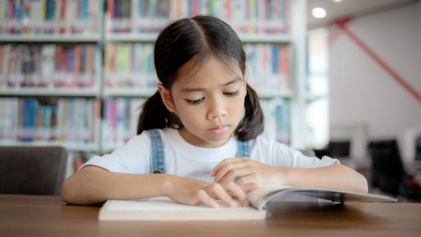 istock A young girl is sitting at a table in a library, reading a book 2153750428