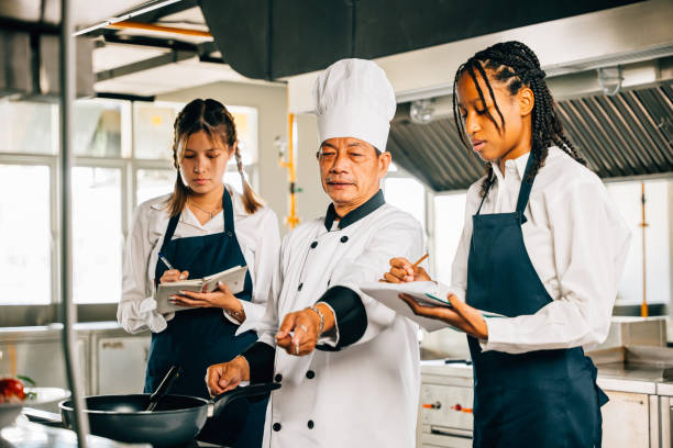 istock Asian senior chef in uniform teaches cooking techniques to multiracial students. Emphasizing teamwork learning and note-taking in this professional kitchen workshop. Food Edocation 2153756838
