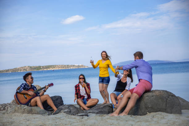 Group of happy young friends playing guitar, listening music, drinking beers on sandy beach party in sunny summer day against sea and cloudy blue sky. There are three girls and two boys sitting on rocks.