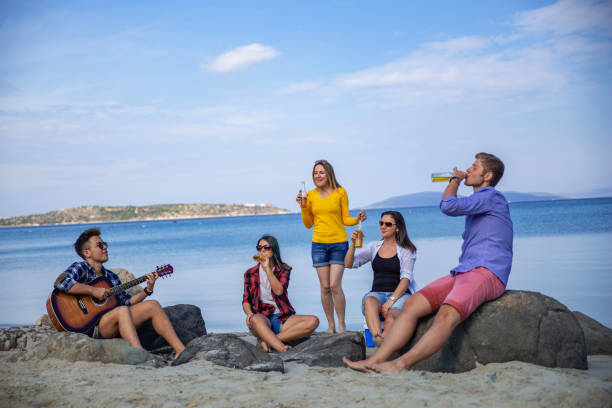 Group of happy young friends playing guitar, listening music, drinking beers on sandy beach party in sunny summer day against sea and cloudy blue sky. There are three girls and two boys sitting on rocks.