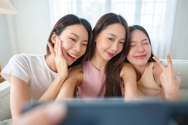 istock Three young Asian women smiling and making peace signs while taking a selfie with smartphone on a couch, capturing a joyful and friendly moment indoors. 2153763366