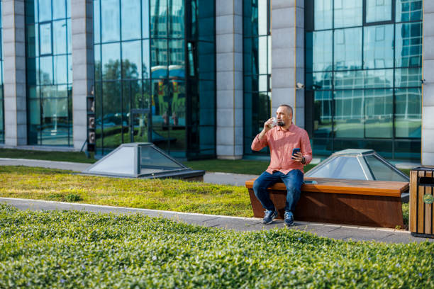 A mature Caucasian man sitting comfortably on a bench, focused on his mobile phone, in a serene and pleasant outdoor setting