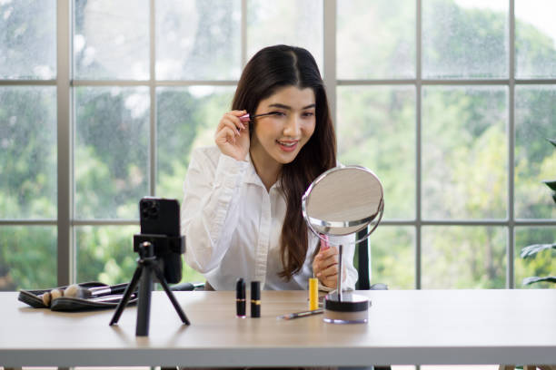 istock Young asian woman is applying makeup while looking into a round mirror on a table, recording a makeup tutorial with a smartphone. 2153786245