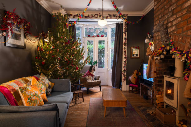 A wide shot of a cosy living room decorated for Christmas in a home in South Shields, North East England. The wood-burning stove is lit, there is a large Christmas tree in one corner and there is a pet dog sitting on an armchair wearing a patterned jumper and green sparkly costume reindeer antlers.Videos similar to this scenario available.