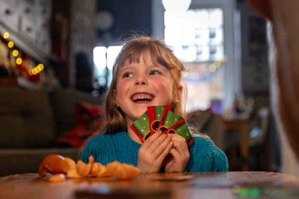 A low-angle shot of a young girl smiling and holding a fan of playing cards, looking away from the camera. There is a partially peeled tangerine on the table in front of her and she is sitting in her home in South Shields, North East England. In the background the kitchen is visible out of focus.Video similar to this scenario available.