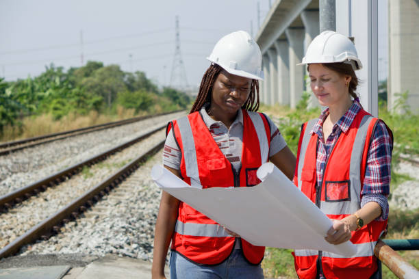 istock Two construction or engineer with safety helmet and high-visibility vest reviewing work to be done or progress on a railway track project while holding a set of plan or blueprint. 2153799963