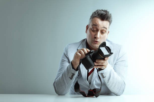 Middle-aged man in a light gray blazer inspects a professional camera with a focused expression, dark gray shirt underneath, sitting at a white surface with a simple background