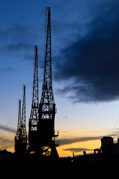 This image features large cranes at a port, with a clear sky and a distant cityscape in the background. The cranes, showing signs of wear, are equipped with long jibs and hoists, indicative of heavy lifting operations. The foreground reveals a section of the dock with various shipping equipment, hinting at the industrial nature of the location. The photograph captures the essence of maritime logistics and the infrastructure necessary for handling large cargo vessels.