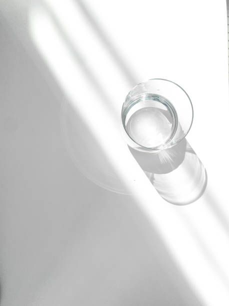 A minimalist stock photo featuring a clear glass of water casting a soft shadow on a white surface. Sunlight streams diagonally across the scene, creating a calm and serene atmosphere. Ideal for themes of purity, simplicity, hydration, and wellness.