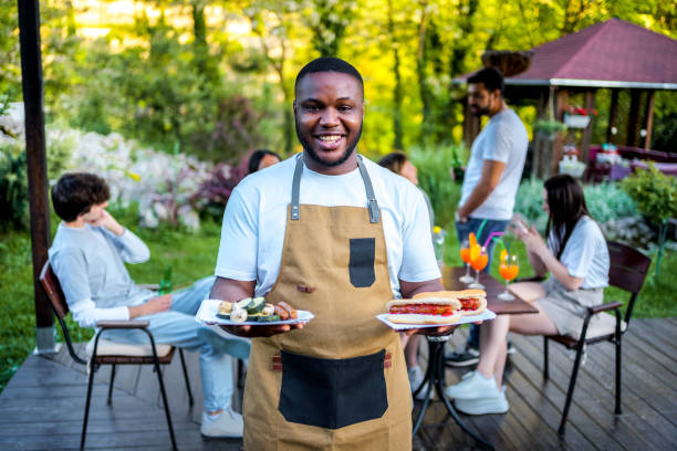 Portrait of a young cheerful man holding food during a backyard barbecue party with his friends behind