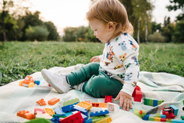 Kid 2 year old play constructor game sitting in green grass on blanket in park sunny summer day. Outdoors creative activities. Toddler boy little cute walking and playing small car toys in garden. niño de 2 años juega al juego de constructor sentado en la hierba verde sobre una manta en el parque día soleado de verano. actividades creativas al aire libre. niño pequeño lindo caminando y jugando juguetes pequeños de coche en el jardín.